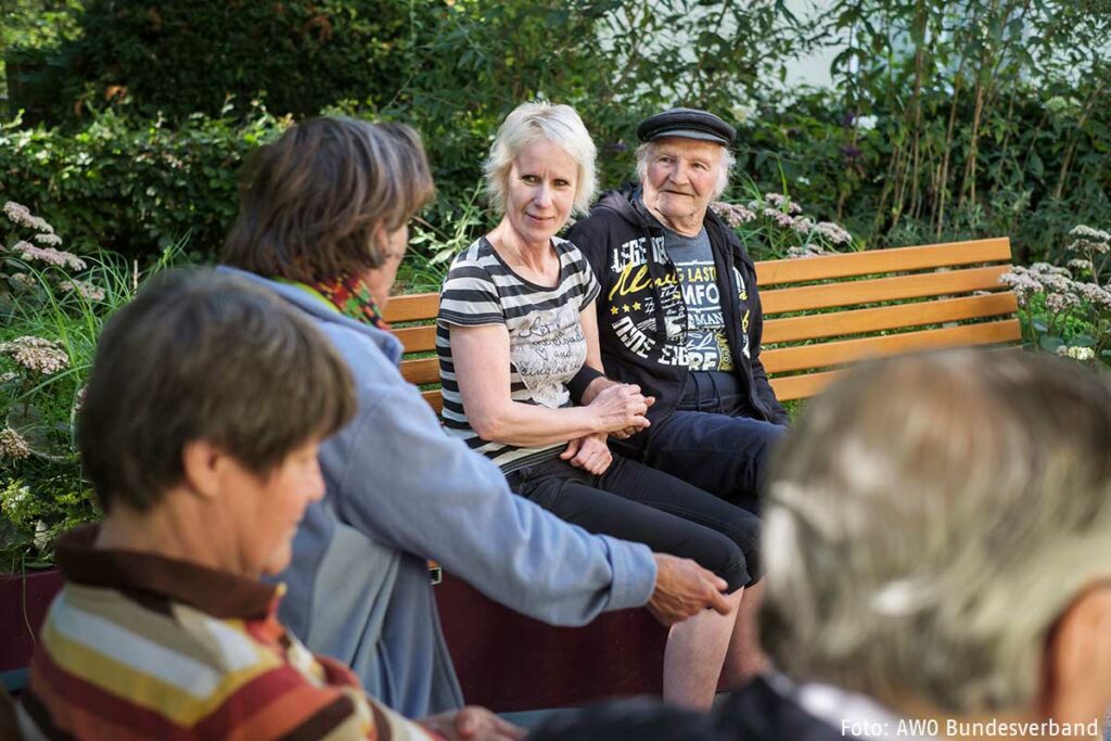 Mehrere alte Menschen Senioren und Seniorinnen sitzen draußen auf Bänken und sprechen miteinander und einer Pflegekraft