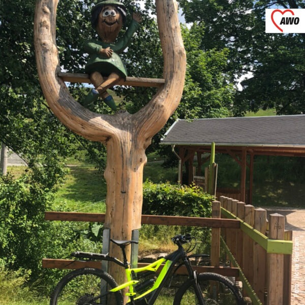 ein neongelbes Fahrrad lehnt an einem Baum, in dessen Gabelung eine Holzhexe sitzt