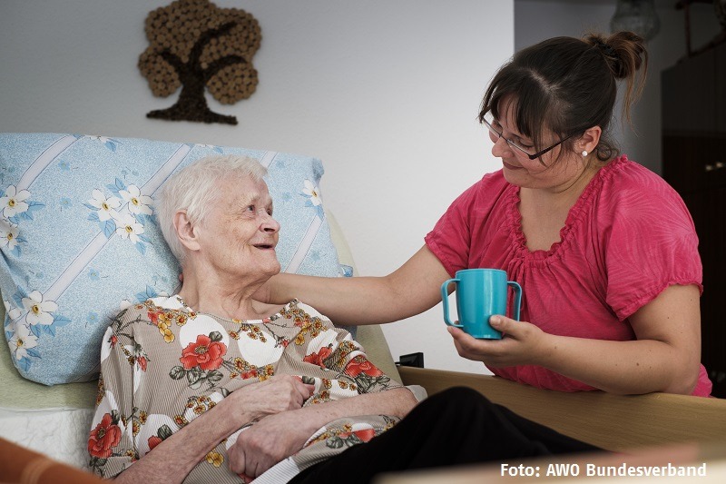 Eine Pflegerin reicht einer alten Frau, die im Bett liegt, eine Tasse Tee