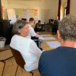Kathrin Sonnenholzner sitzt mit einer Gruppe Freiwilliger in einem Stuhlkreis im Seminarraum. Im Hintergrund sind Moderationsplakate mit Themen und Notizen sichtbar, einige Freiwillige diskutieren und schreiben auf große Papierbögen am Boden.