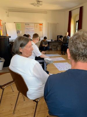 Kathrin Sonnenholzner sitzt mit einer Gruppe Freiwilliger in einem Stuhlkreis im Seminarraum. Im Hintergrund sind Moderationsplakate mit Themen und Notizen sichtbar, einige Freiwillige diskutieren und schreiben auf große Papierbögen am Boden.
