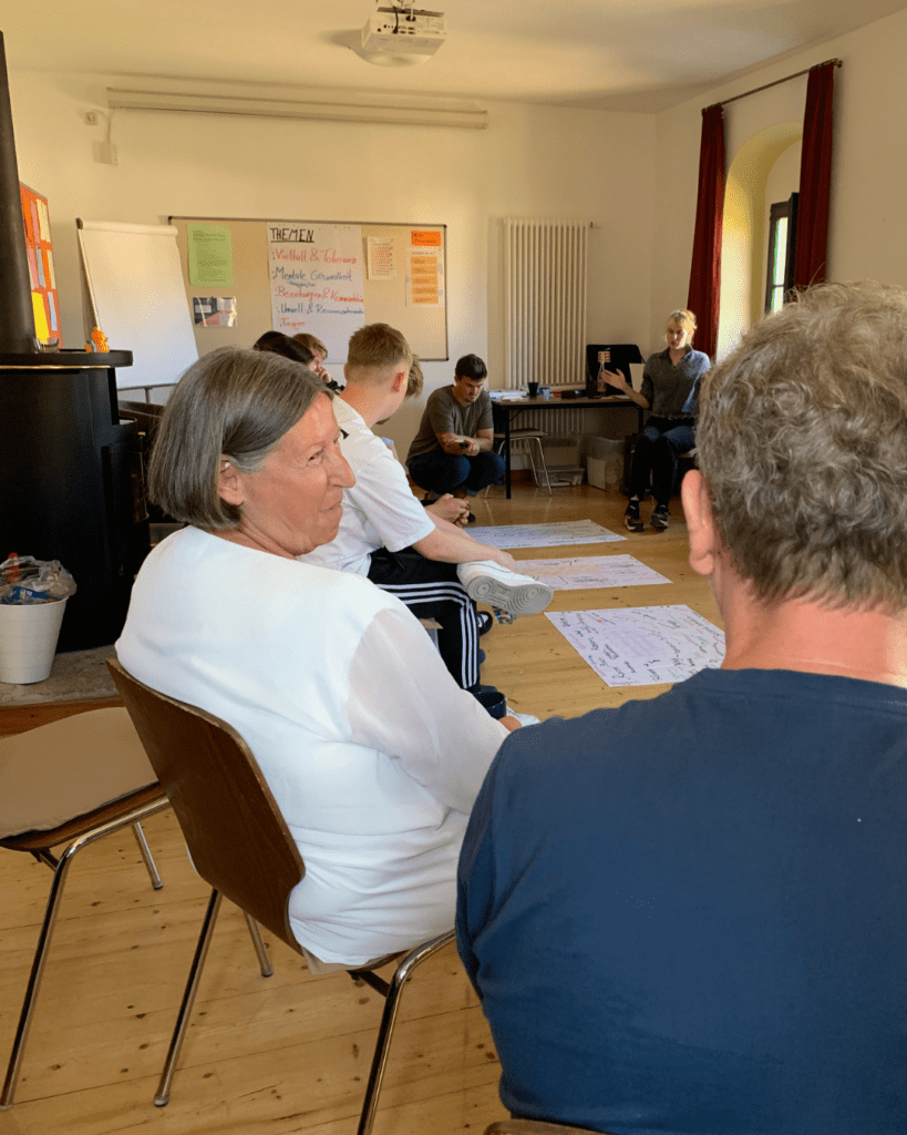 Kathrin Sonnenholzner sitzt mit einer Gruppe Freiwilliger in einem Stuhlkreis im Seminarraum. Im Hintergrund sind Moderationsplakate mit Themen und Notizen sichtbar, einige Freiwillige diskutieren und schreiben auf große Papierbögen am Boden.