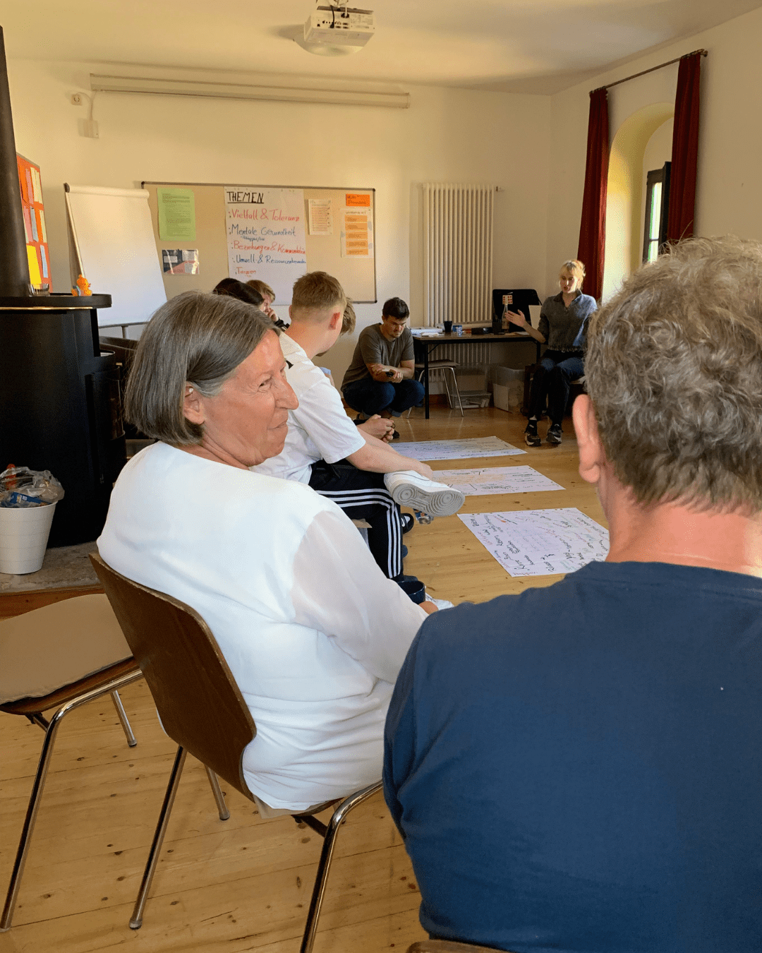 Kathrin Sonnenholzner sitzt mit einer Gruppe Freiwilliger in einem Stuhlkreis im Seminarraum. Im Hintergrund sind Moderationsplakate mit Themen und Notizen sichtbar, einige Freiwillige diskutieren und schreiben auf große Papierbögen am Boden.