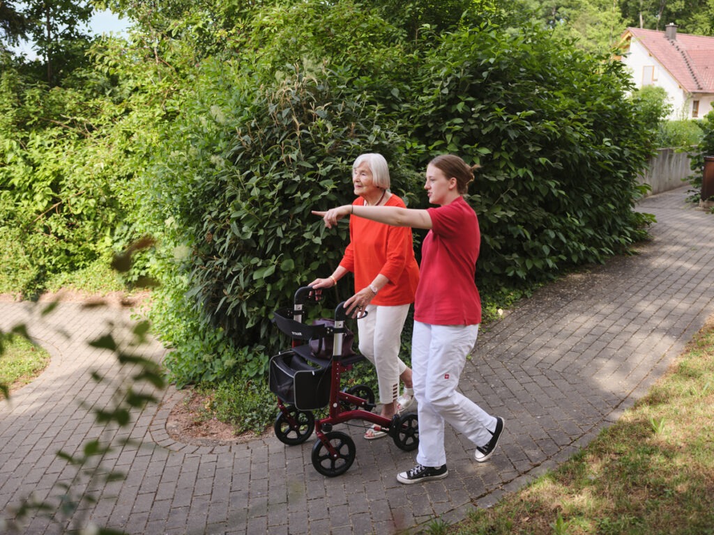 eine junge Frau geht mit einer älteren Frau im Rollator im Park spazieren