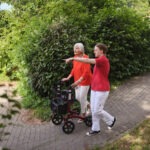 eine junge Frau geht mit einer älteren Frau im Rollator im Park spazieren