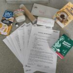 Auf einem grauen Boden liegen die Materialien einer AWO-Seminarbox „Mit allen Sinnen“. Zu sehen sind mehrere ausgedruckte Anleitungen, verschiedene Lebensmittel wie Salzkaramell-Kokosmilch, Kokosöl, Zitronensäure, Speisestärke und Natron sowie weitere Bastelmaterialien.